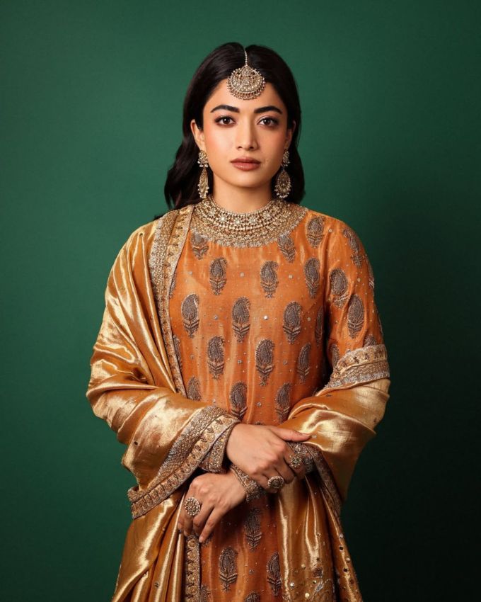 <strong>રશ્મિકા મંદાના :</strong> ડાર્ક ઓરેન્જ અને ગોલ્ડન કલર ટ્રેડિશનલ હેવી ફૂલ સ્લીવ ડ્રેસ પહેર્યો છે જેમાં ફલોસી સિલ્ક ઇન્ટરિકેટ વર્ક ની બોર્ડર વાળો દુપટ્ટો પસંદ કર્યો છે, આખા ડ્રેસમાં છૂટી છૂટી સરખી કેરી ડિઝાઇન જોવા મળે છે, આ ઉપરાંત નેકમાં થોડું ગોલ્ડન વર્ક જોવા મળે છે.